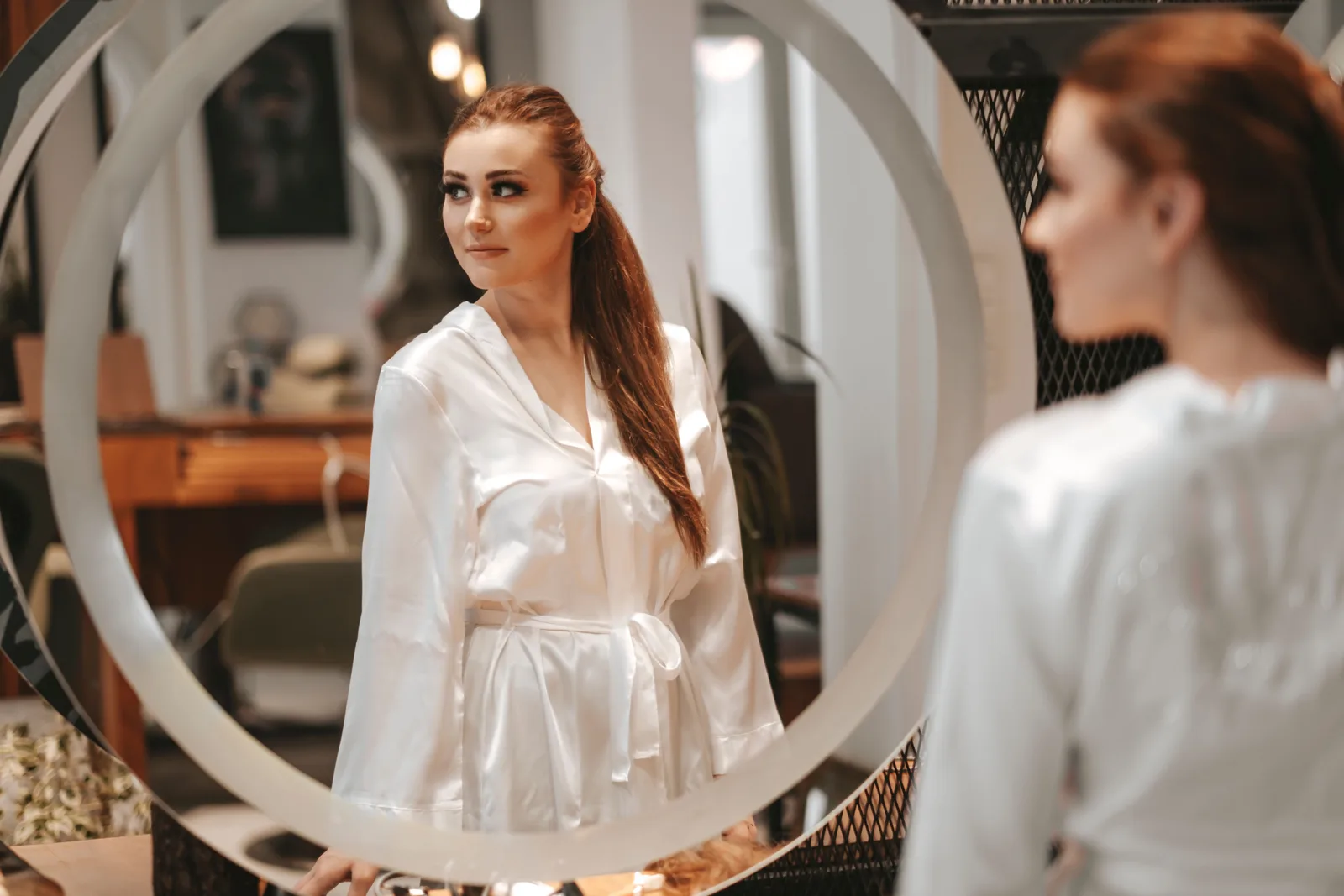 A woman in a white robe looking at her reflection in a round mirror