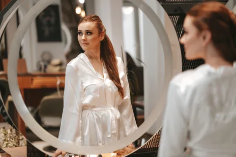 A woman in a white robe looking at her reflection in a round mirror